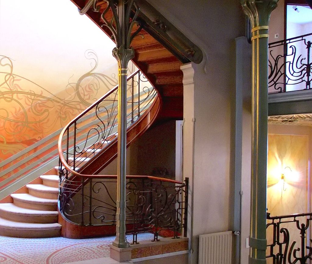 A stair curves between levels beside slender columns, patterned walls and flowing iron railings.