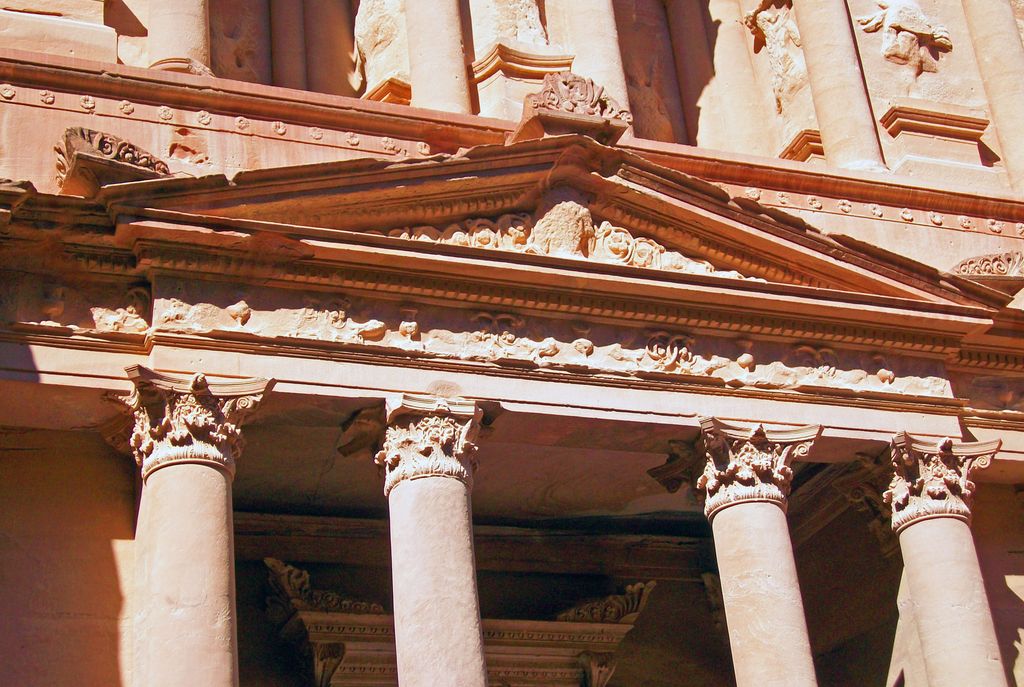 A triangular rock-cut pediment with deep mouldings and sculpture stands below tall columns and divided upper crowning forms in warm red stone.