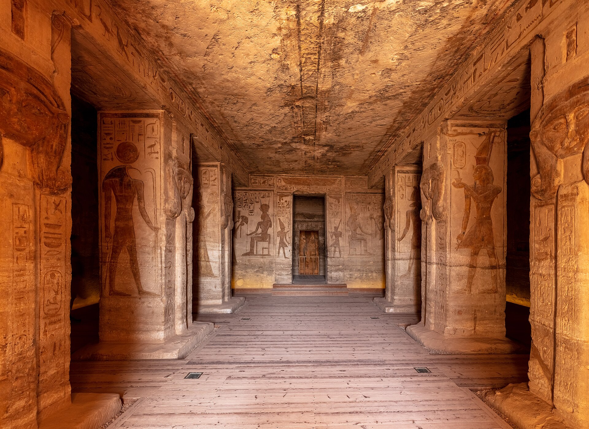 Small Temple interior looking between Hathor-headed pillars along a timber-floored passage to the carved vestibule wall and rear doorway.