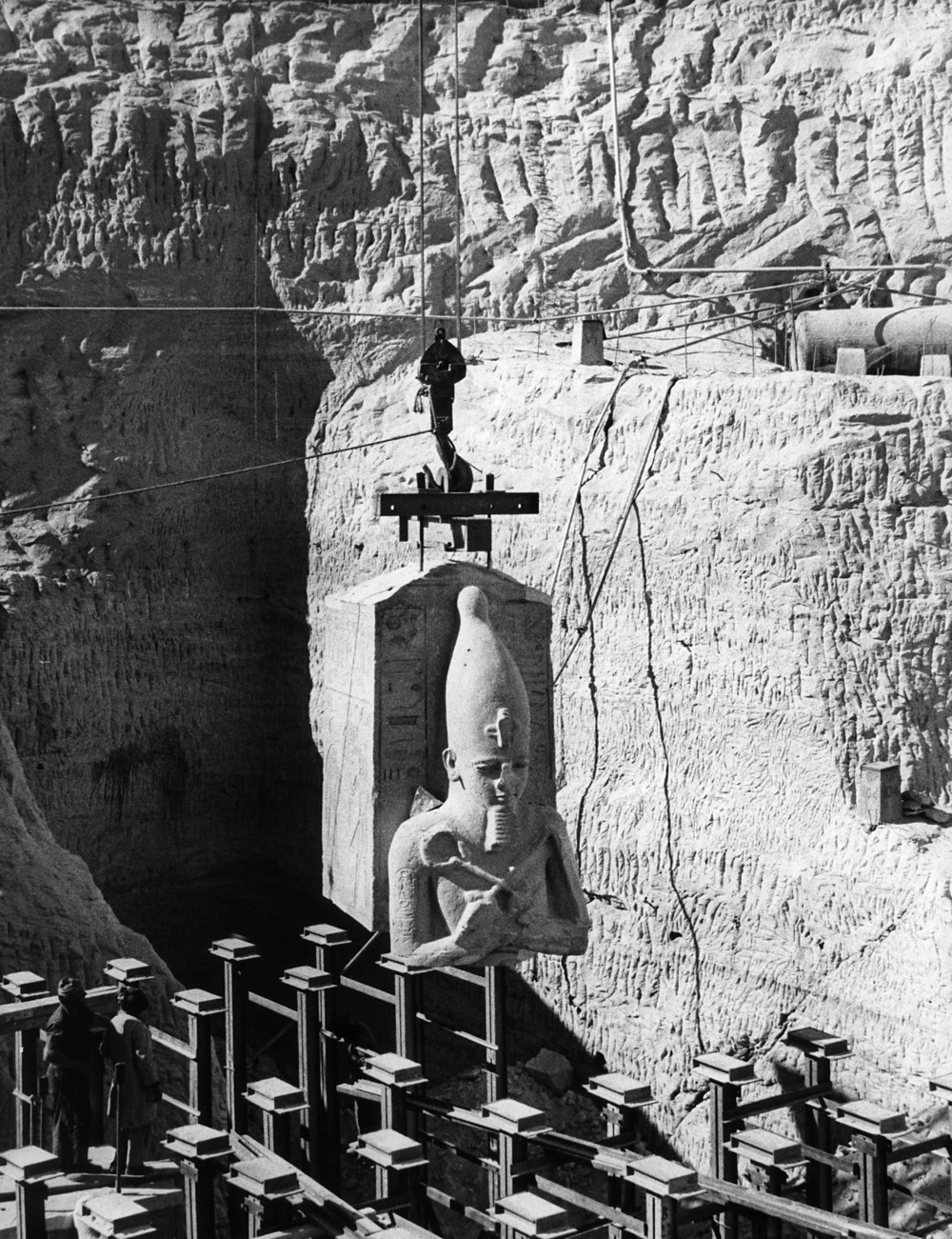 Black-and-white salvage photograph of an Osiride king pillar block being suspended on lifting equipment over a cut temple wall.