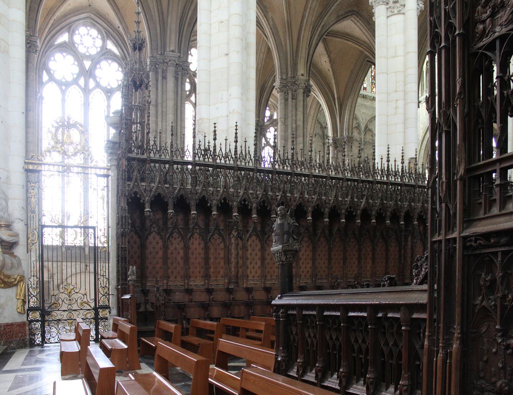 Dark carved wooden choir stalls with tall Gothic cresting stand behind metal gates and modern benches beneath pale stone piers, pointed arches and bright windows.