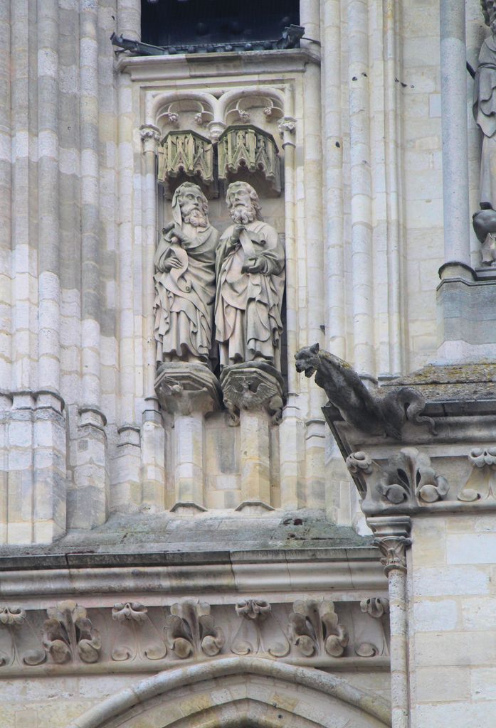 Two carved stone figures stand beneath small canopies among clustered facade shafts, beside foliage ornament and a projecting animal-shaped waterspout.
