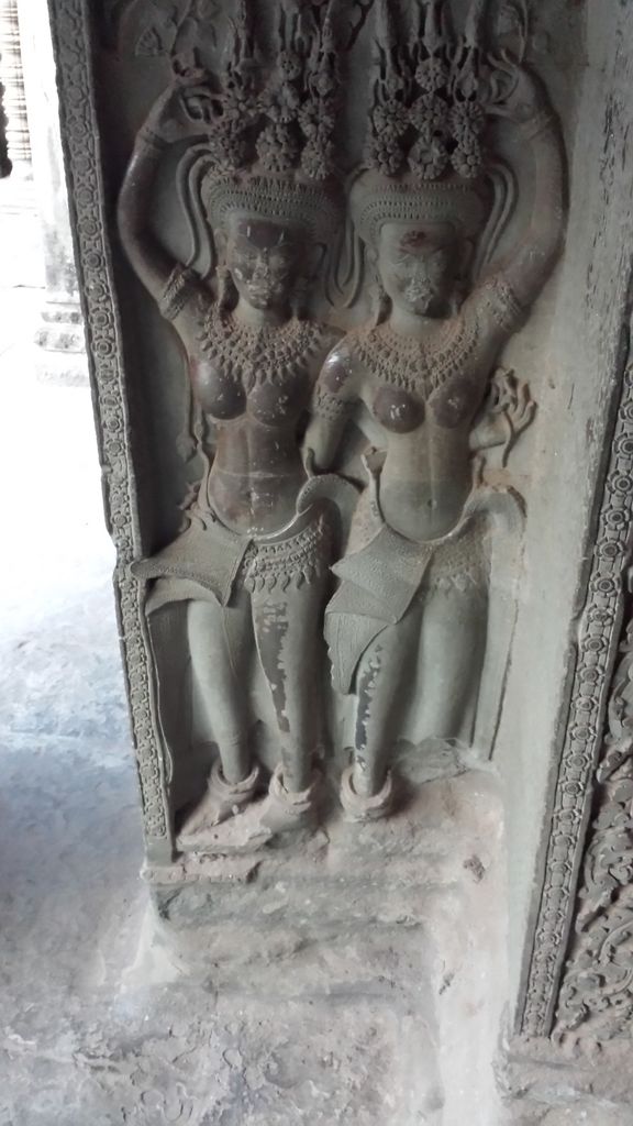Two standing female figures are carved in shallow relief with elaborate headdresses, necklaces, patterned skirts and different raised-hand gestures.