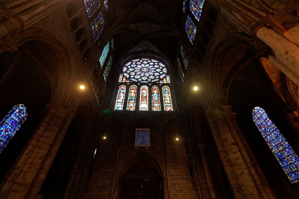 A luminous stained-glass rose and five tall lancets shine in a dark cathedral transept, with other coloured windows, stone arches and vaults surrounding them.