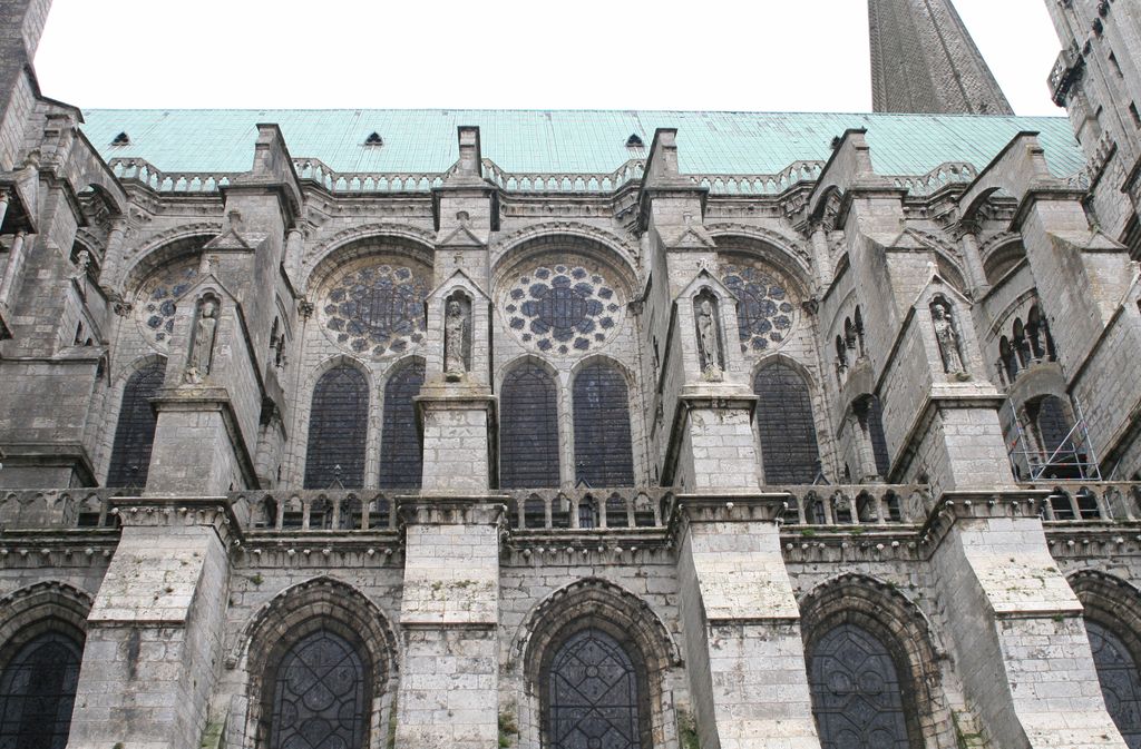 A frontal exterior view shows repeated tall windows and small roses between heavy stone buttress piers topped by figures, with roof and partial flying arches above.
