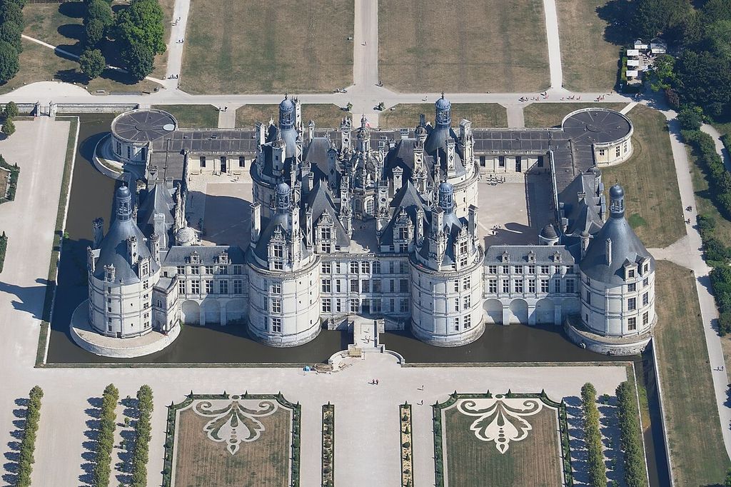 An aerial view shows a square white-stone château with four round towers, dense roof turrets, surrounding courts, moat and formal gardens.