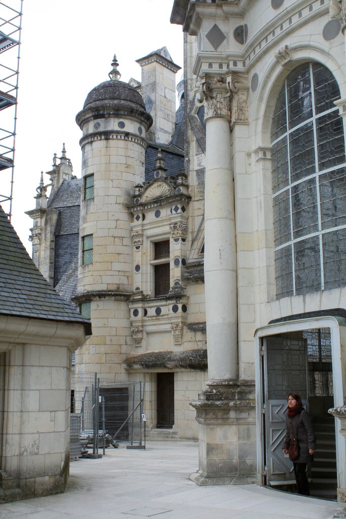 A roof terrace passes among pale carved stone turrets, chimneys, steep slate roofs and a large arched window, with scaffolding at the left.