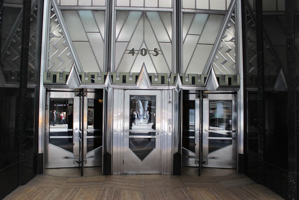 Three silver metal and glass entrance doors sit beneath stepped geometric frames, frosted glass, chevrons and the number 405 between polished black walls.