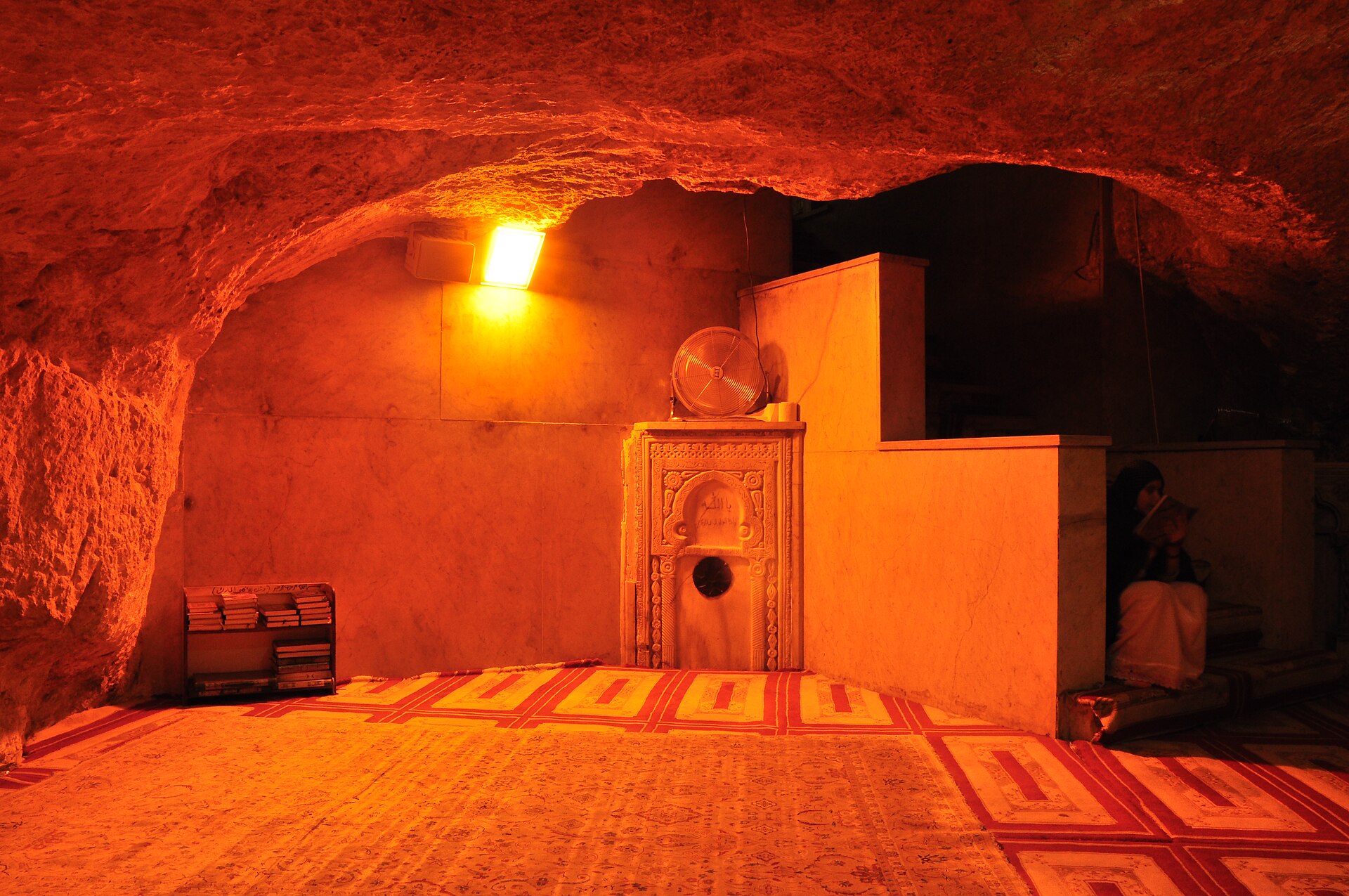 Cave beneath the rock with a present prayer niche, matting, lamps and visitors.