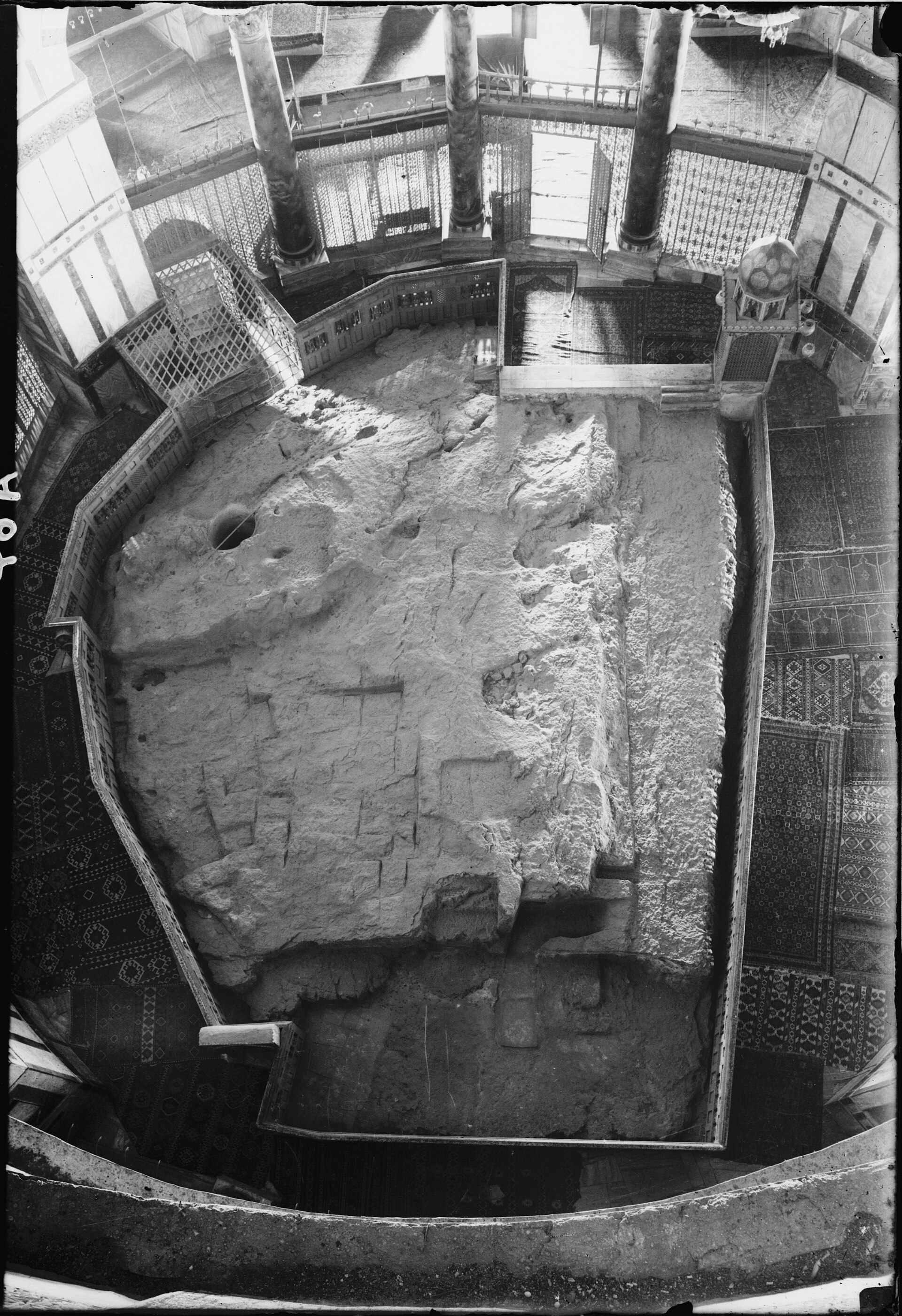 Historic high view of the irregular exposed central rock encircled by a barrier, columns and patterned carpets.