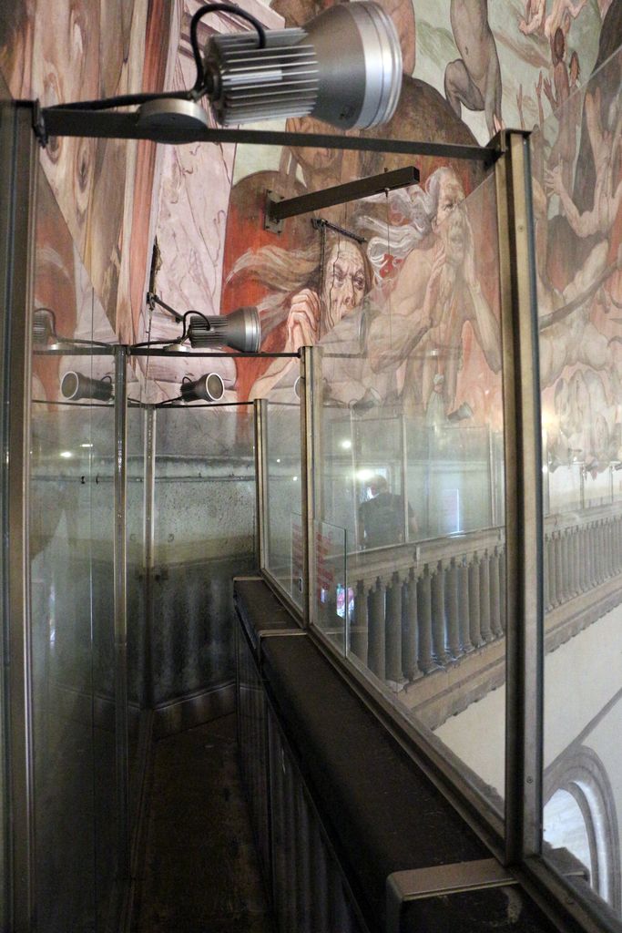 A narrow gallery beside a painted curved wall has a stone balustrade, tall glass safety screens and rows of modern spotlights.