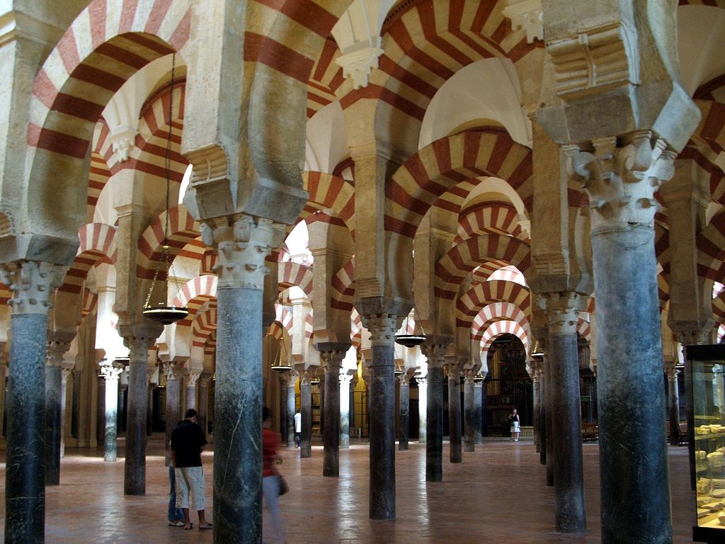 Rows of slender columns support red-and-white striped lower horseshoe arches and upper round arches across a deep interior with visitors and later fittings.