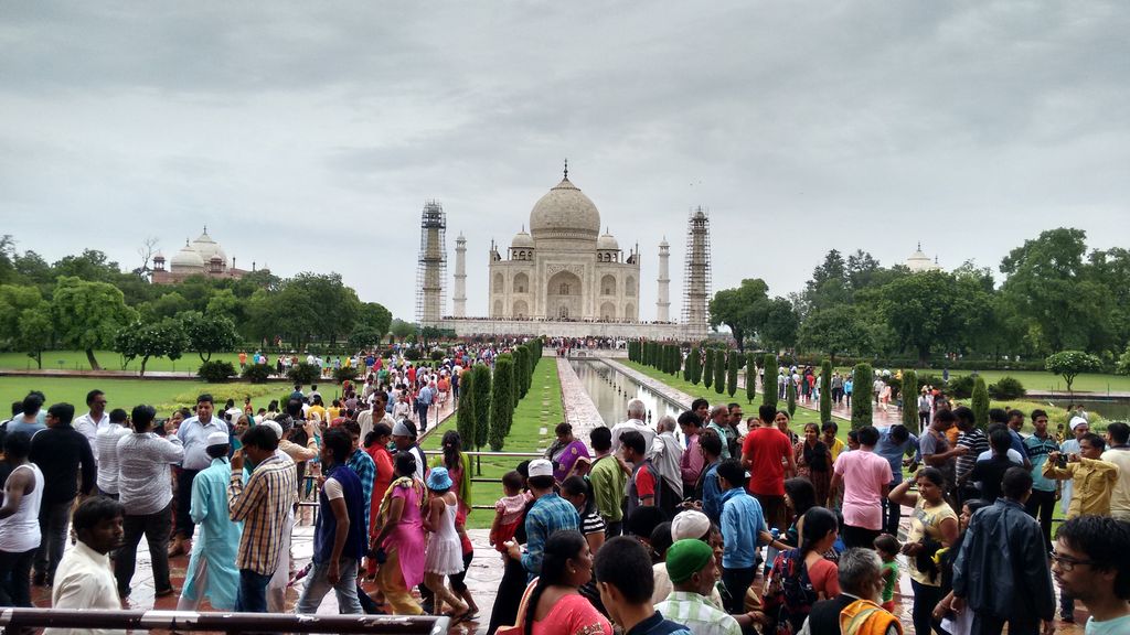The white domed Taj Mahal and four minarets stand at the end of a formal garden and water channel packed with visitors beneath an overcast sky.