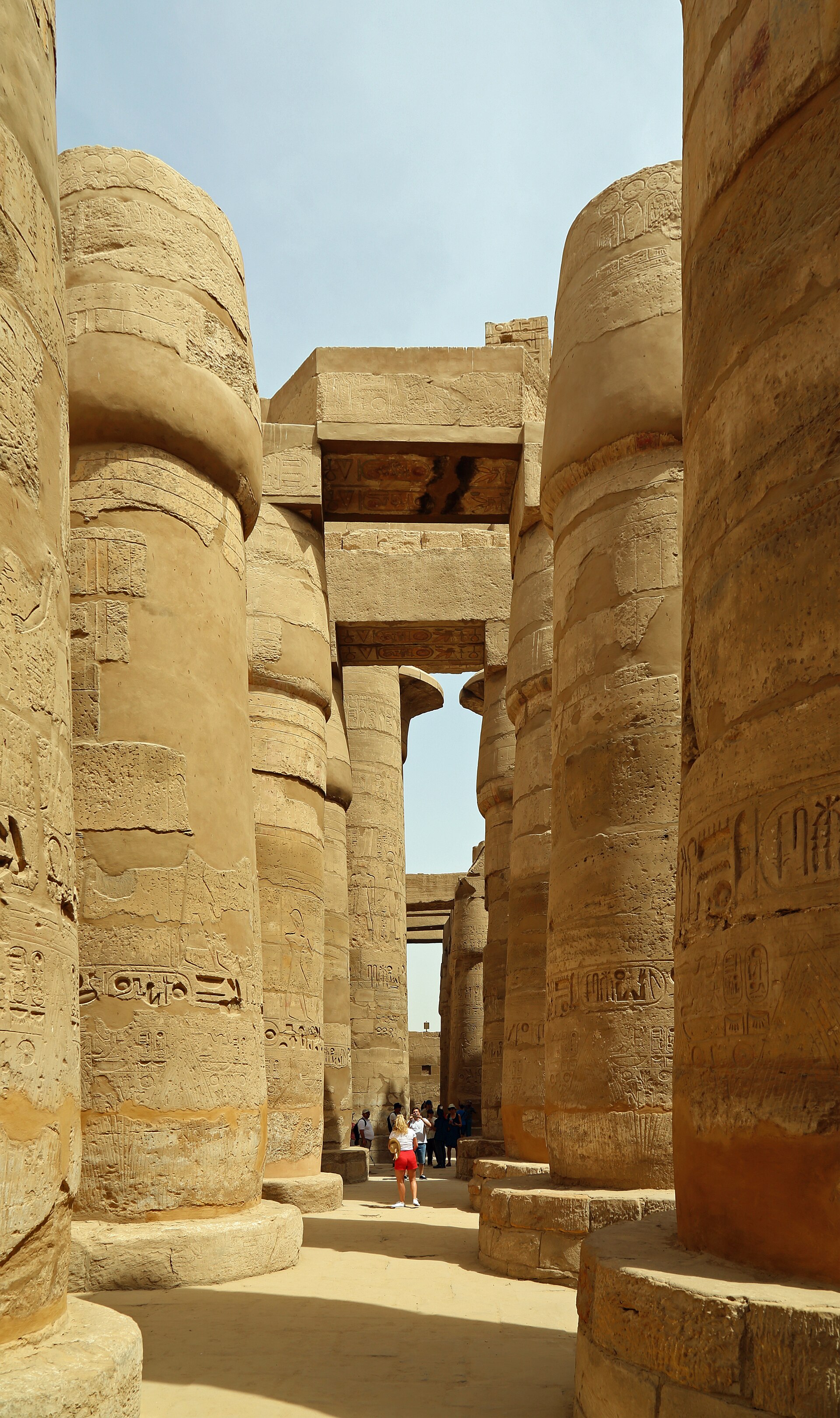 Looking between giant papyrus-shaped sandstone columns towards surviving stone architraves and open sky in the Great Hypostyle Hall.