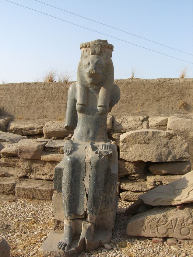 Weathered seated lioness-headed goddess statue within the Mut temple ruins, with missing limbs and a mudbrick wall behind.