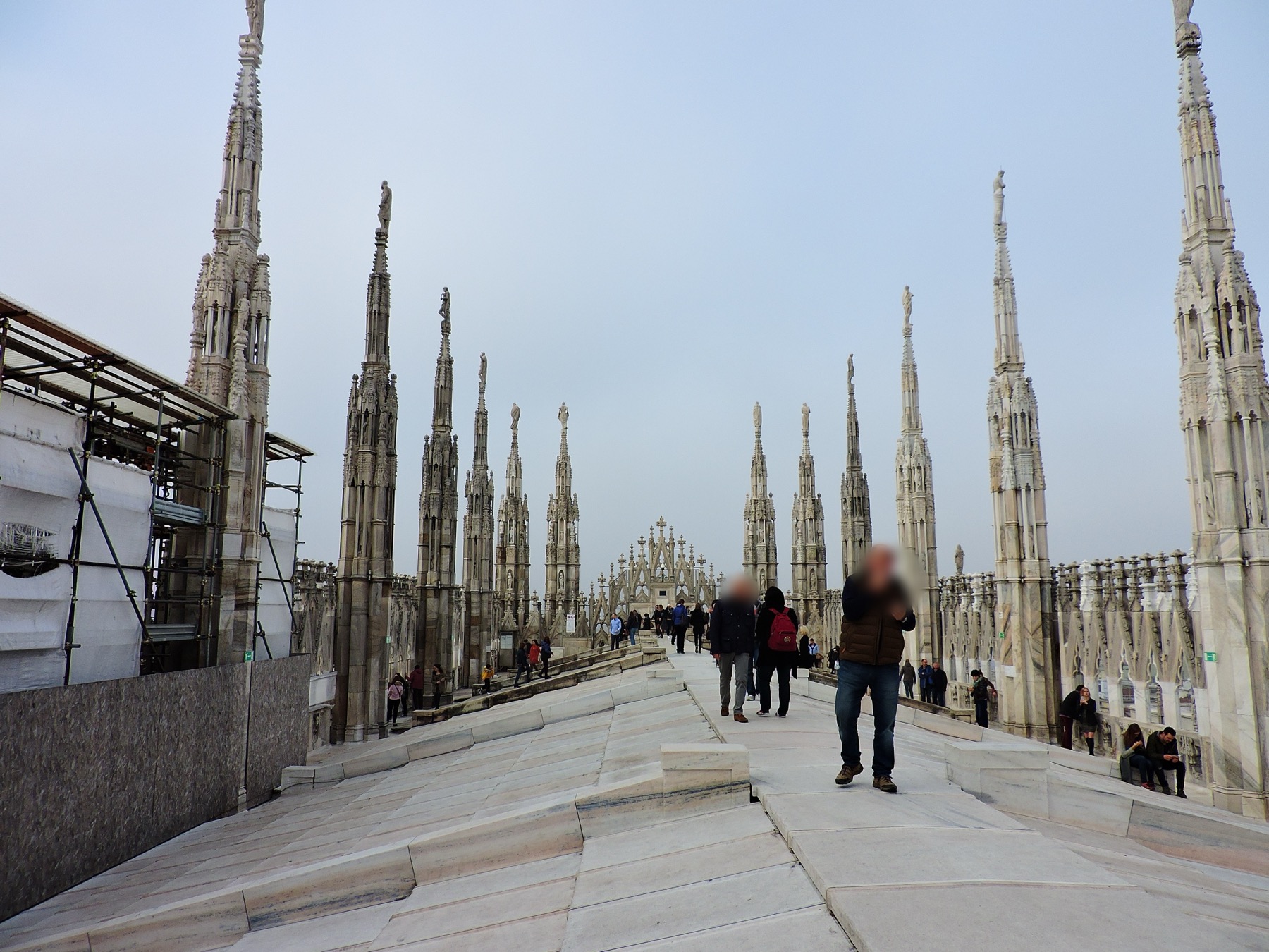 Visitors walk across Milan Cathedral’s sloping marble roof terraces among pinnacles, statues and an active scaffold enclosure.