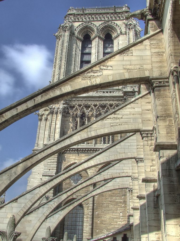 Several long stone flying arches cross diagonally toward a tall cathedral wall, with masonry joints, tracery and part of a tower visible behind.