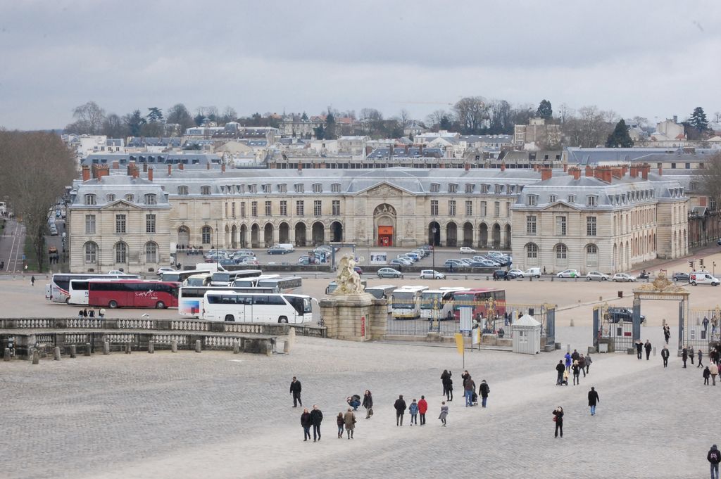 A long pale-stone stable building with arcades and a central pediment stands across a broad square filled with buses, cars, gates and pedestrians.