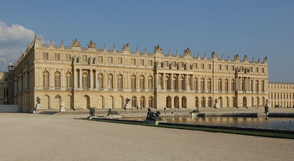 A long pale-stone palace facade with repeated arched windows, columns and roof statues stands beside a reflecting pool and gravel terrace.