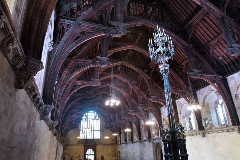 Dark timber hammerbeam trusses and curved braces repeat above a vast pale-stone hall, with hanging electric lights and a tall metal lamp standard.