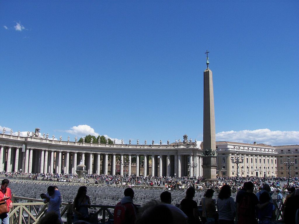 A long curving colonnade lined with roof statues bends around an obelisk and a crowded square filled with rows of temporary seats.