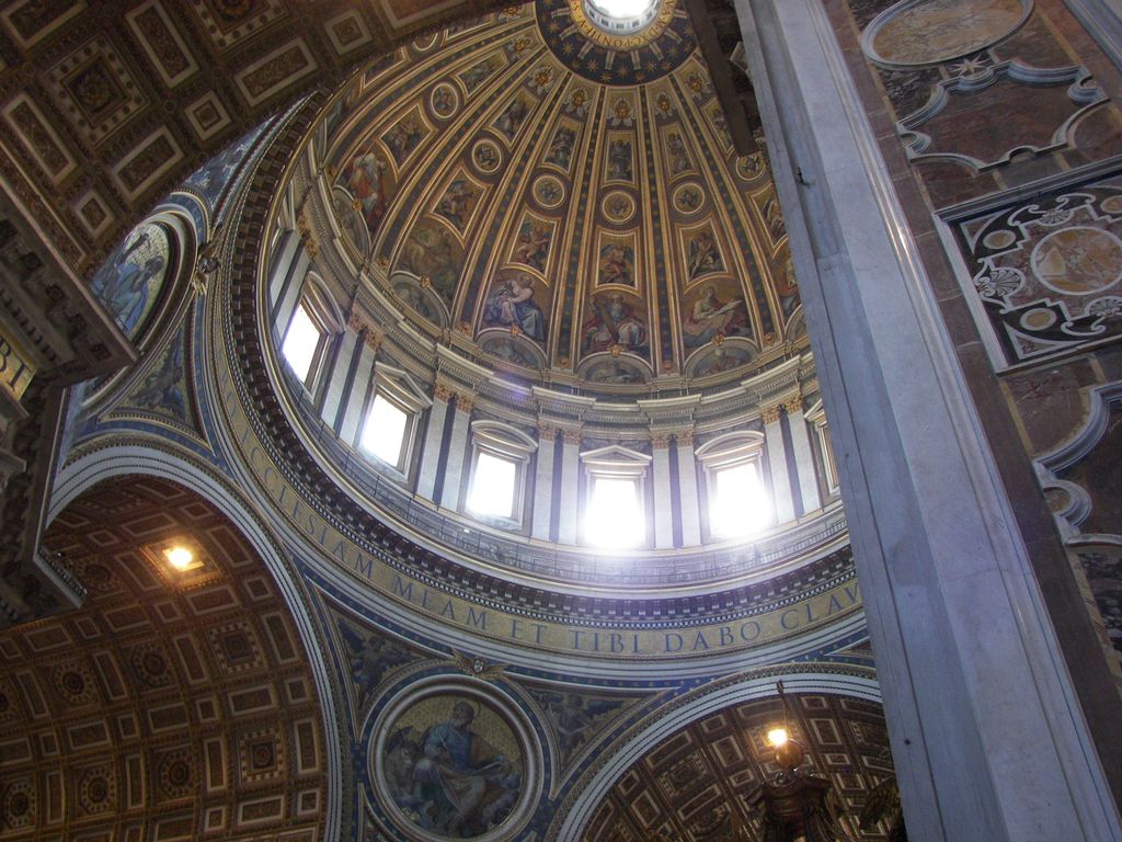 An ornate dome interior rises above bright drum windows, an inscribed ring and painted or mosaic panels, partly blocked by a massive stone pier.