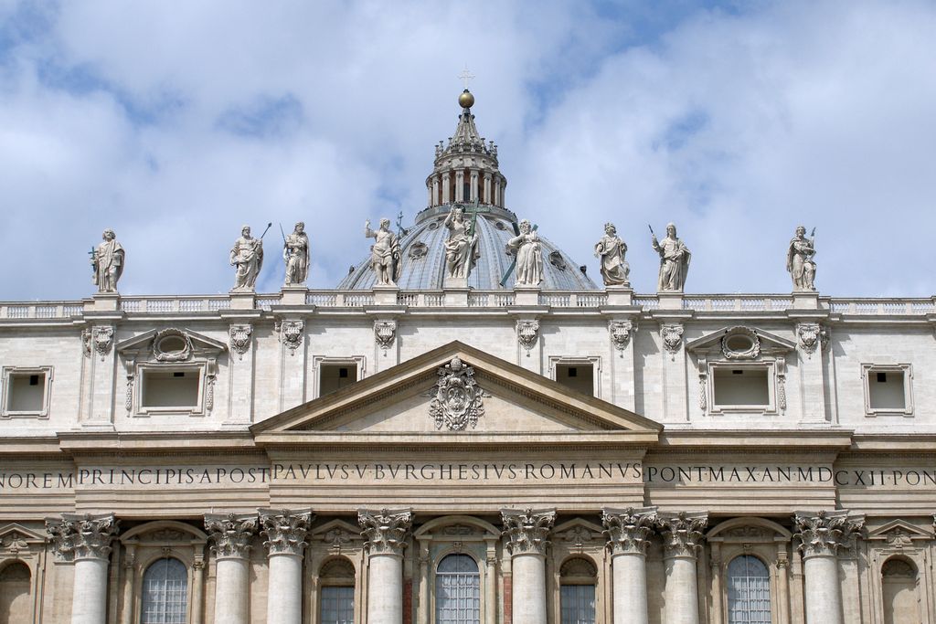 The upper centre of St Peter's broad classical facade has giant Corinthian columns, a pediment, long Latin inscription and roofline statues, with a dome lantern behind.