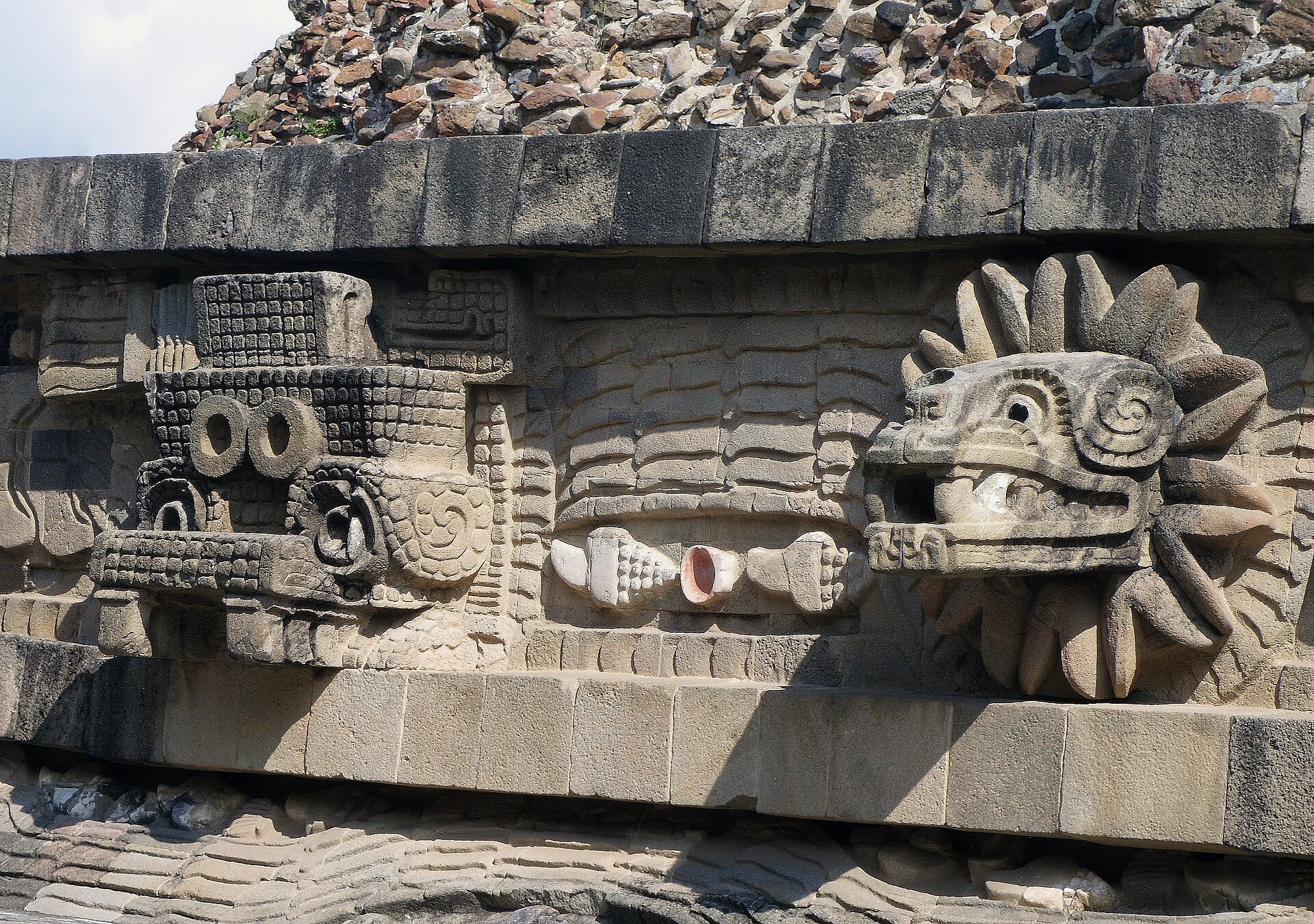 Close view of two different carved projecting heads and linked serpent relief within horizontal stone bands.