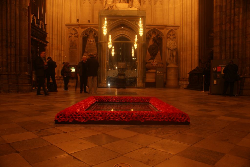 A dark rectangular grave slab surrounded by vivid red poppies lies in the foreground of a warmly lit Gothic nave with visitors beyond.