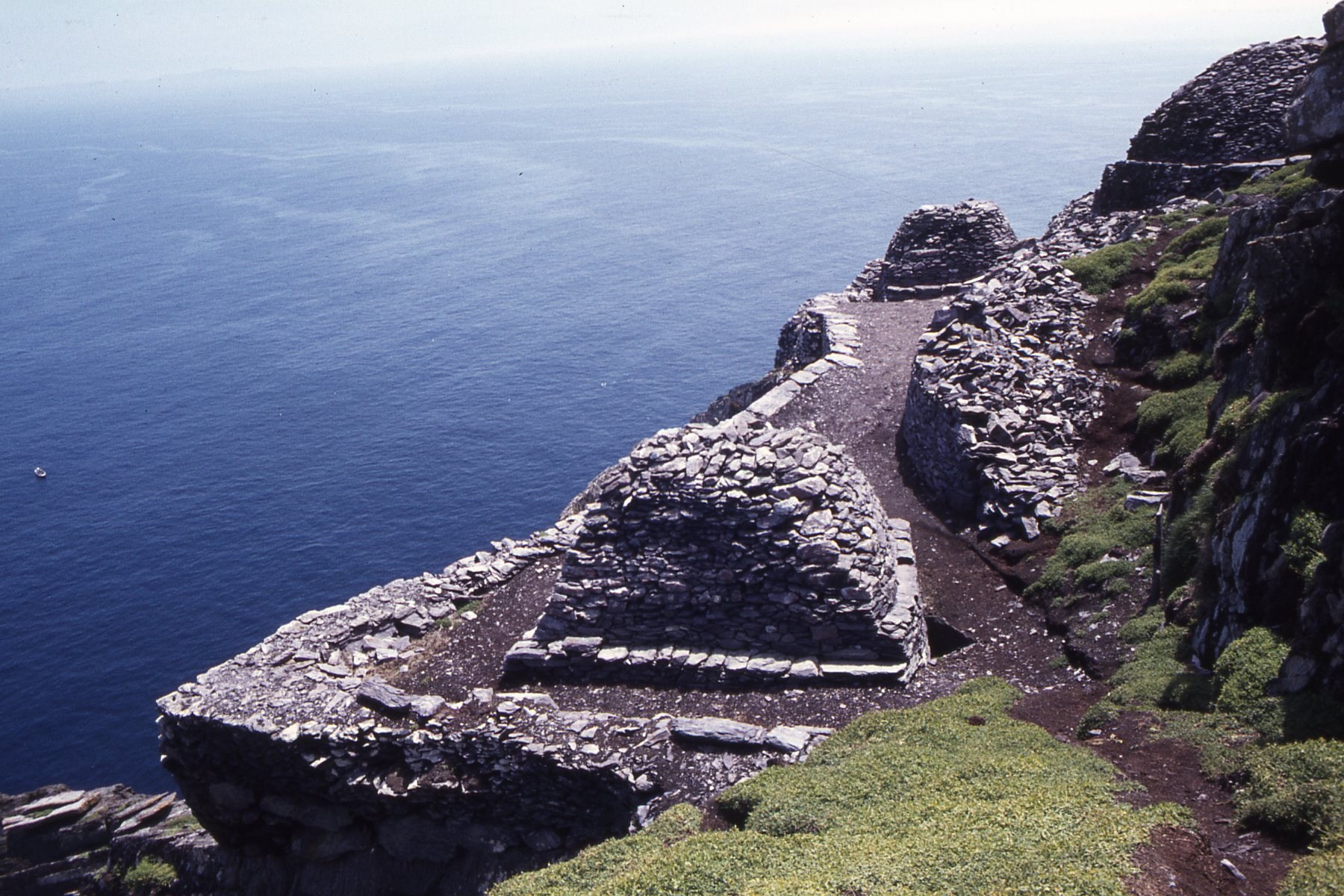 Partly unroofed tapered stone enclosures and terraces below a steep Skellig slope with the sea beyond.