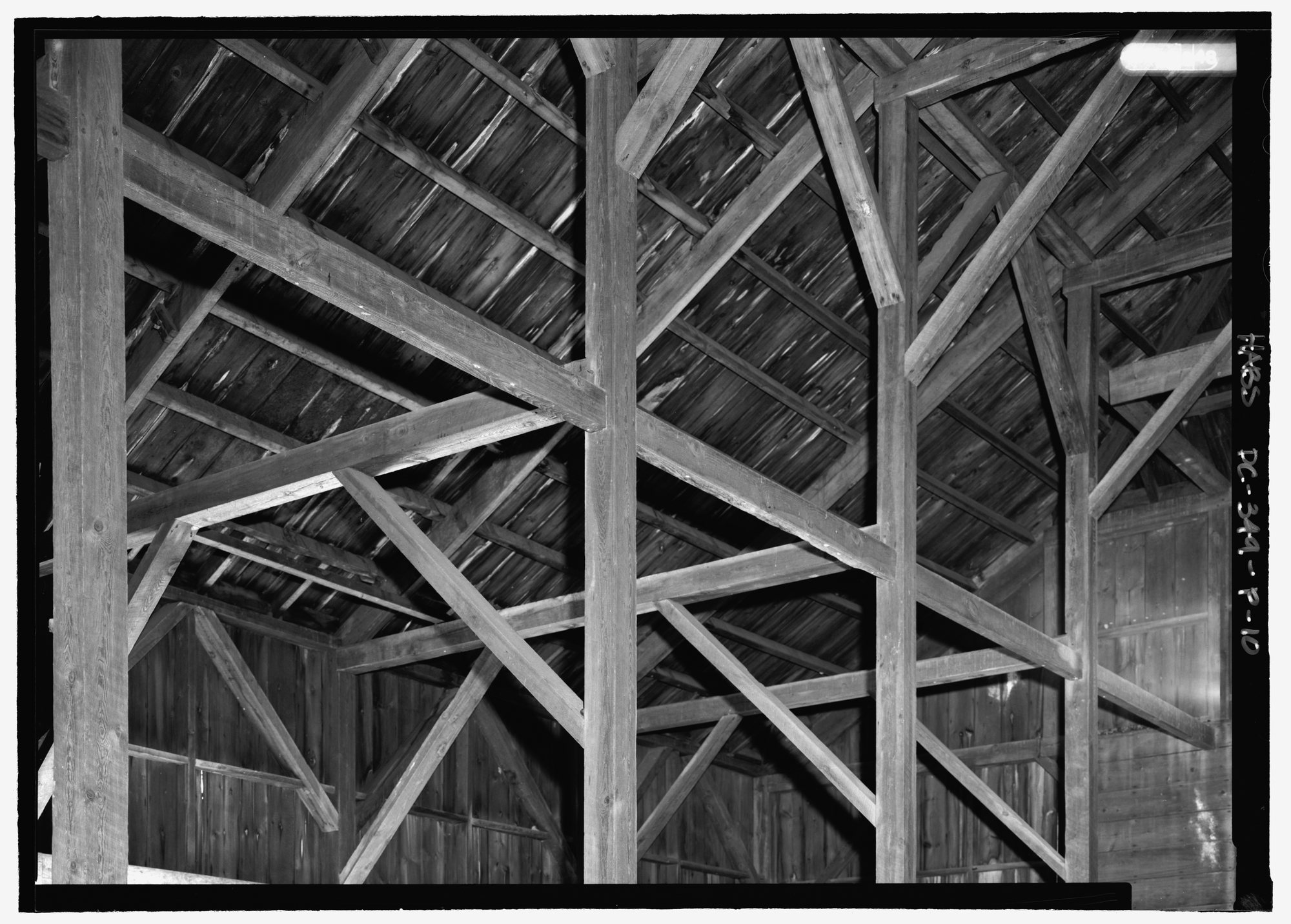Greyscale interior photograph of heavy timber posts, diagonal bracing and roof sheathing at a barn corner.
