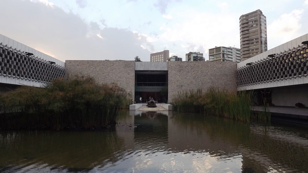 Open-air end of Mexico City anthropology museum courtyard with pond, reeds and galleries on both sides.