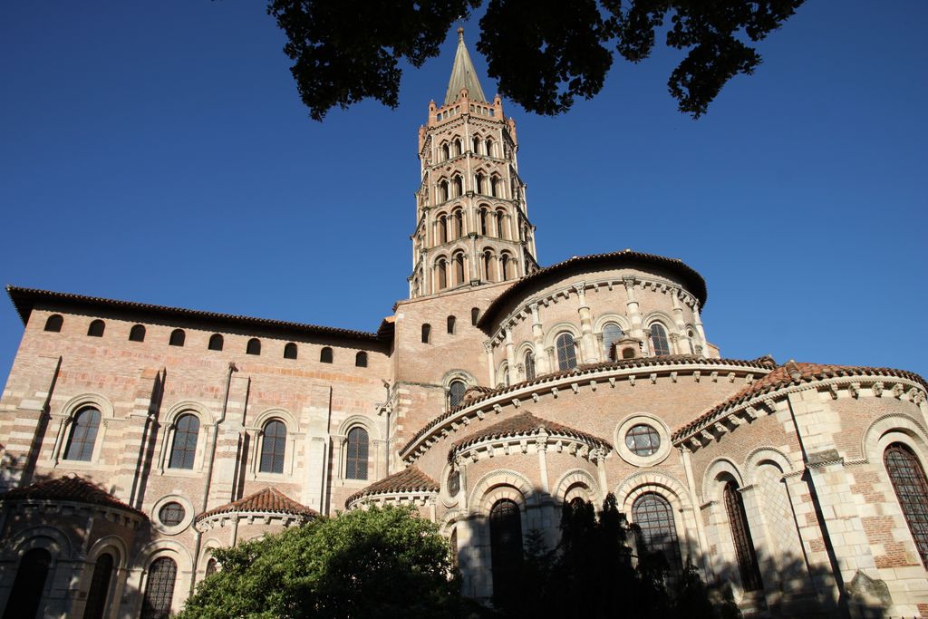 Small curved chapels project below a lower encircling roof and tall apse, with the transept and tiered tower behind.