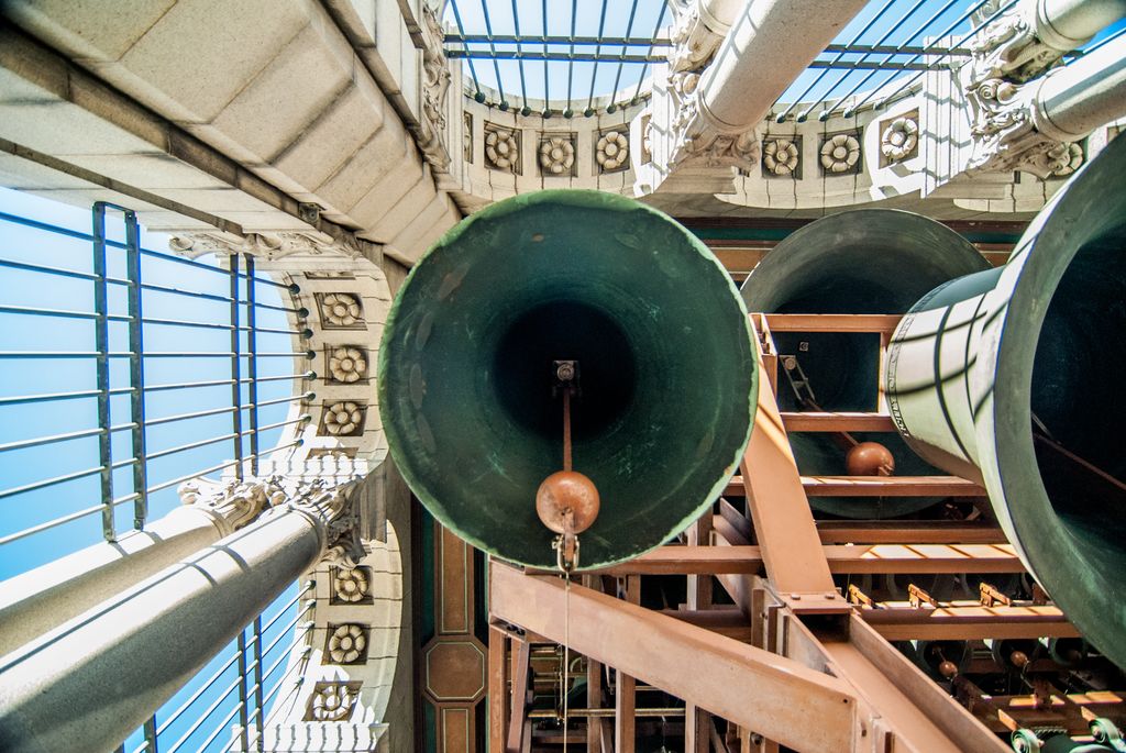 An upward view shows bell mouths and clappers above steel beams, surrounded by the tower’s stone openings and grilles.