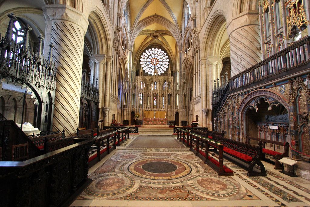 Opposing choir seats and carved canopies stand between patterned stone piers, with an altar and tall stone screen beyond.