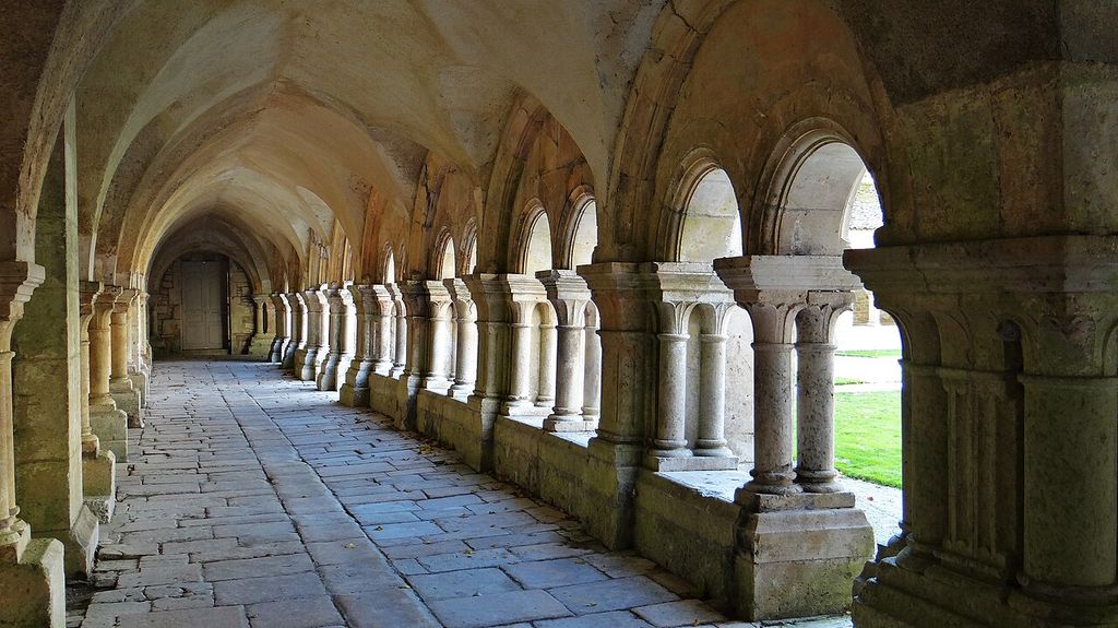 A stone-paved vaulted walk runs beside paired columns and rounded arches opening onto a grassy courtyard.