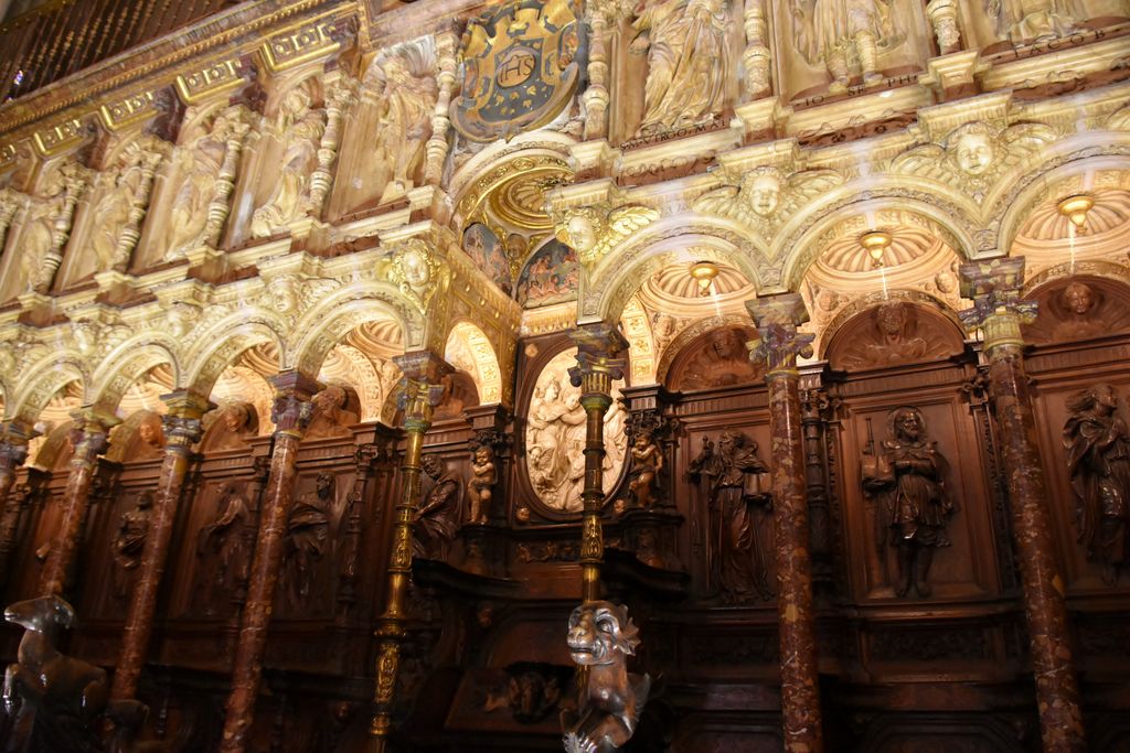 Carved wooden seats and tall relief-covered backs repeat beneath small arches and a sculpted upper register.