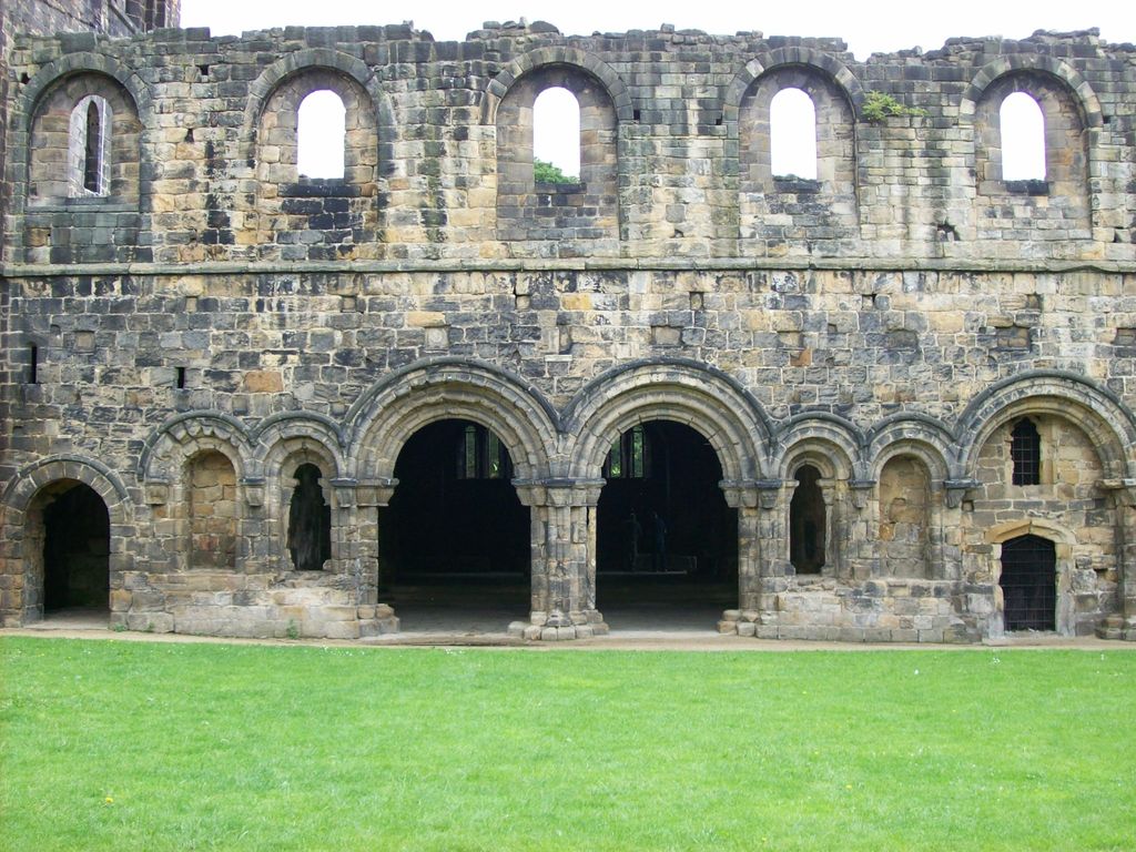 A surviving stone range has paired round-arched entrances, smaller arches and upper openings, facing an open grass foreground.