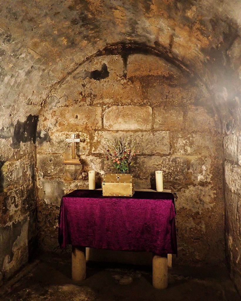 A small altar with purple cloth, candles, flowers and a box stands beneath a low barrel vault between rough stone walls.
