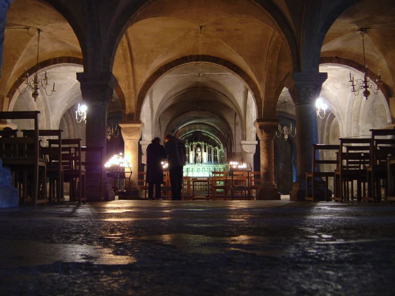 A low stone-floored interior has thick columns and repeated round arches, with chairs, candles and two visitors near a lit altar.