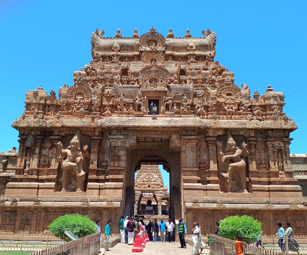 An ornate stone gateway with large guardian figures flanks an open passage, where visitors walk towards buildings beyond.