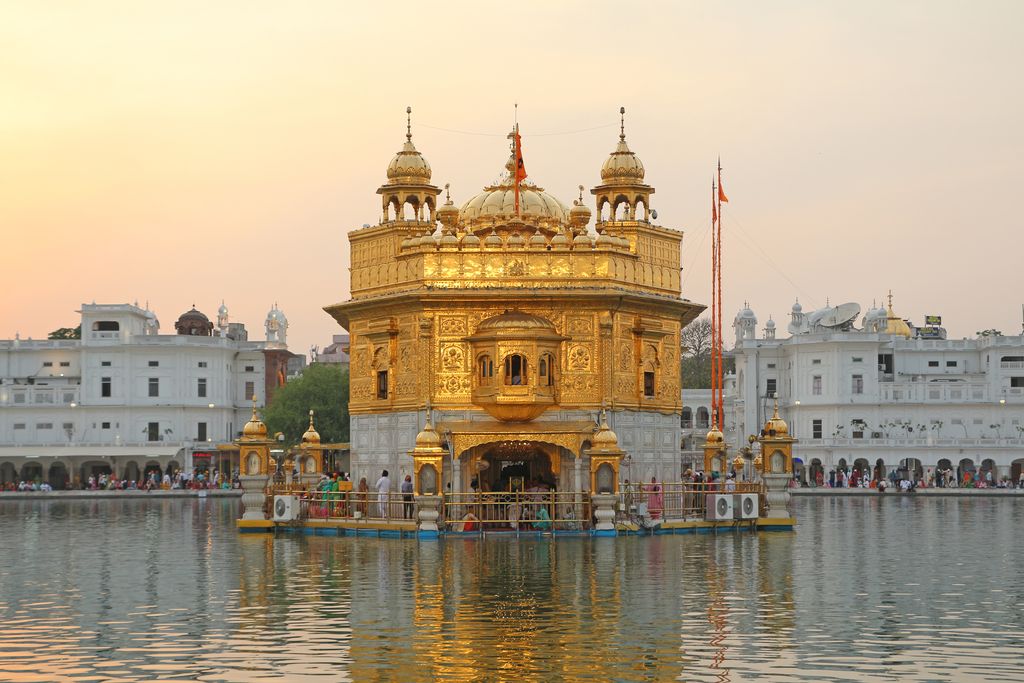 A gilded shrine with a central dome and projecting balcony stands within a pool, with visitors on its surrounding passage and white buildings beyond.