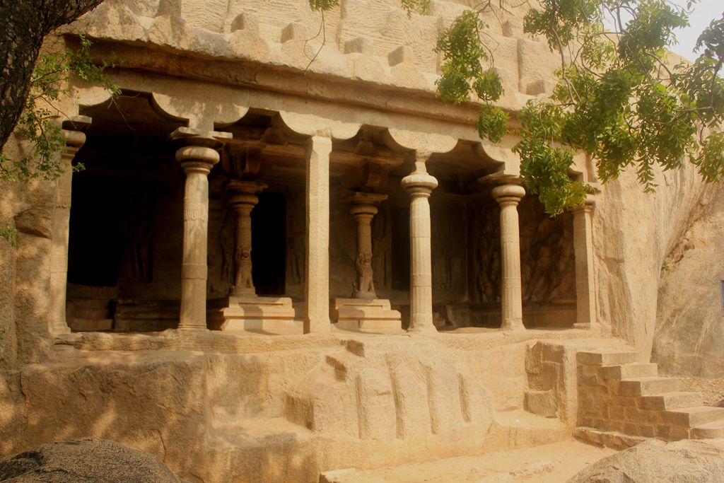 Columns and pilasters frame dark openings cut into a rock face, with lion-based supports inside and steps at the right.