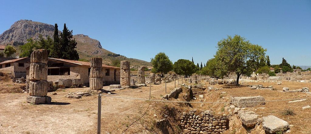 Aligned column drums and excavated foundations of Corinth’s South Stoa, with a rocky hill beyond.