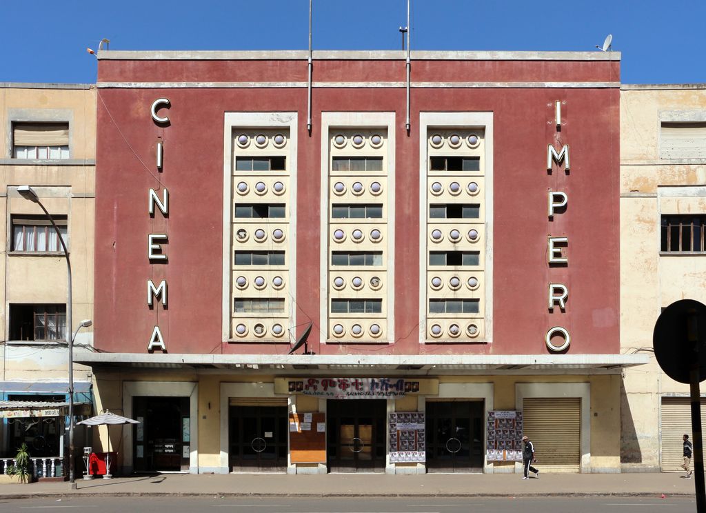 A red cinema frontage has three tall window panels with circular fittings, vertical lettering and a low projecting canopy.