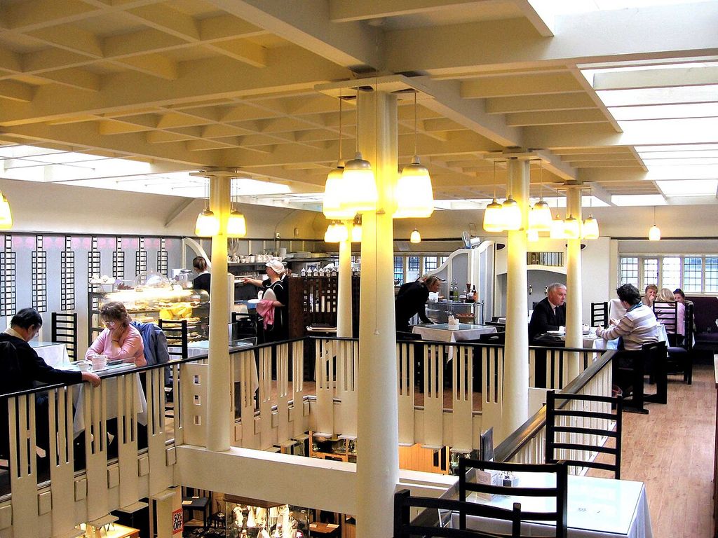 Pale columns and a ceiling grid frame a gallery with vertical railings, tall dark chairs and patterned glazing.