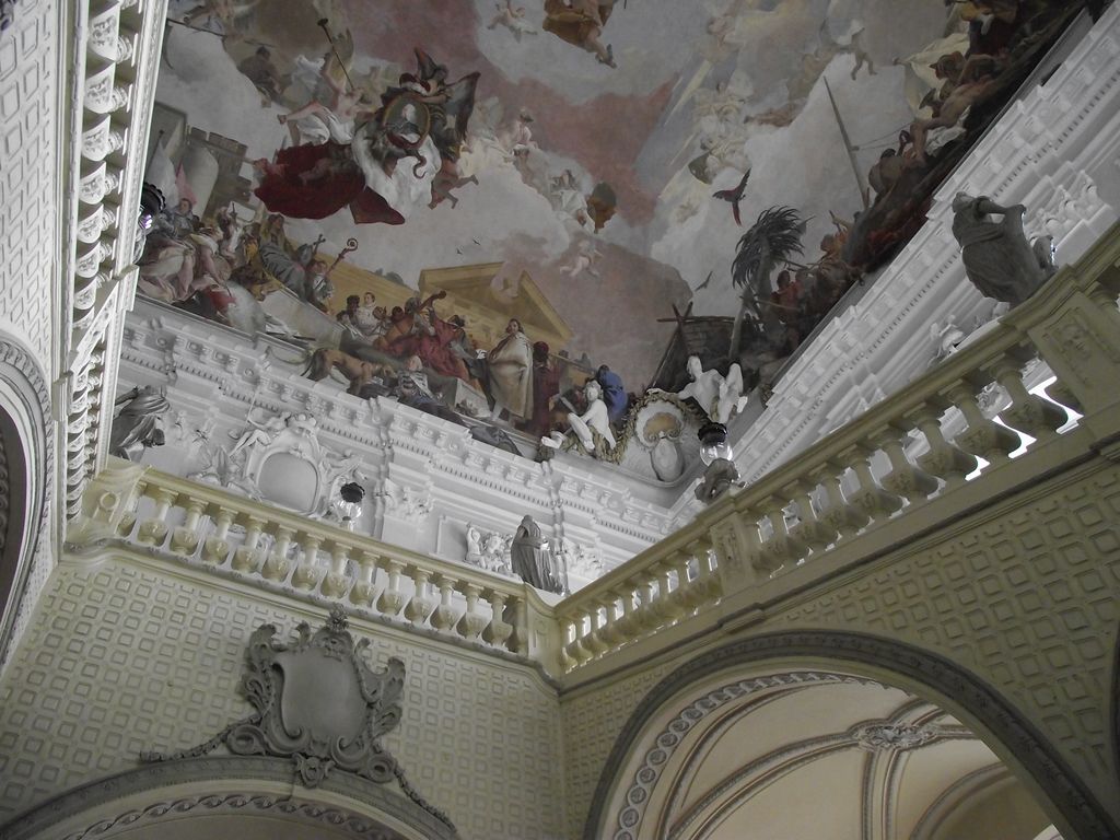 Upward view of stone balustrades and sculpted figures beneath a painted sky in Würzburg Residence’s staircase hall.