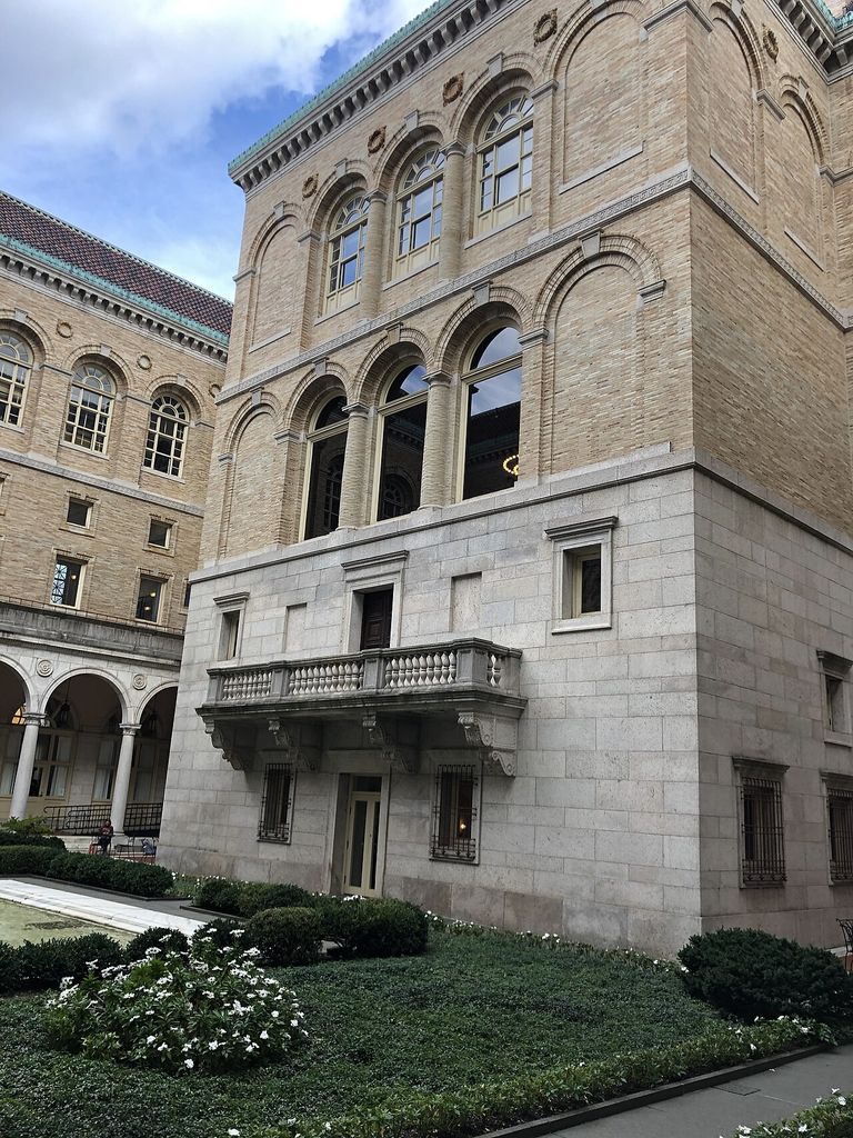 Arched openings and brick upper walls rise above a stone base beside a planted courtyard and arcade.