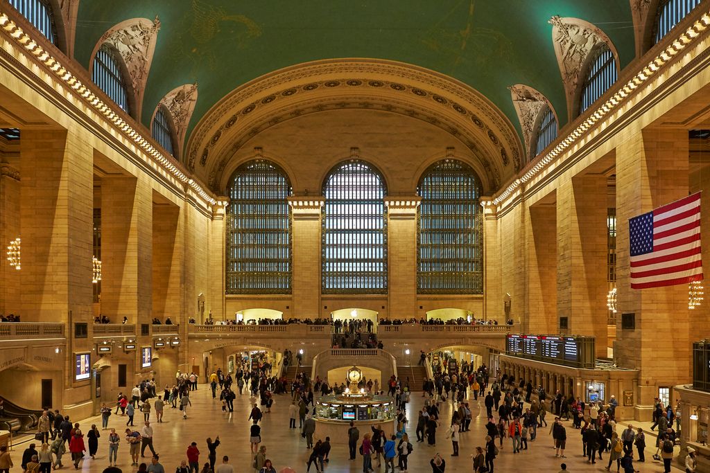 Passengers circulate around a central clock beneath a vaulted ceiling, tall arched windows, galleries and a broad stair.