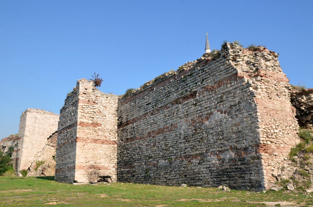 Stone and brick curtain wall between projecting tower remains, with broken wall tops and grass in front.